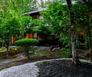 Traditional wooden ryokan building framed by green garden, gravel path and stone lantern in Yamakawa Onsen, Kumamoto