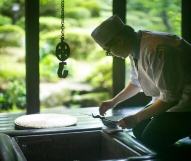 Chef tending a traditional irori hearth in a wooden dining alcove overlooking a moss garden at Wadaya, Ishikawa