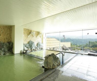 Spacious indoor onsen bath with rock features and large windows overlooking wooded hills at Nunobiki Onsen Komoro Nagano