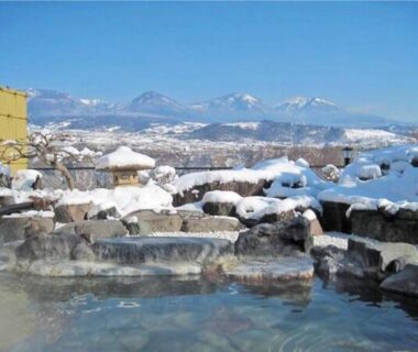 Steaming rock-lined outdoor bath framed by snow and distant mountains at Nunobiki Onsen Komoro, Nagano