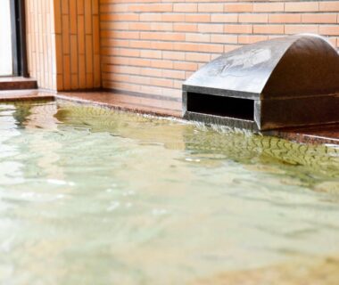 Indoor tiled onsen with metal water spout and brick wall at Shikibu no Yakata Izutsuya Fukushima