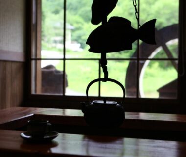 Hanging iron kettle silhouette over wooden table with teacups, wood-framed window opening to green Nagano landscape