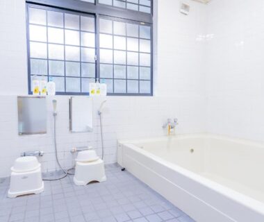 Communal Japanese bathing room with stools, handheld showers, tiled floor and deep soaking tub beneath frosted window