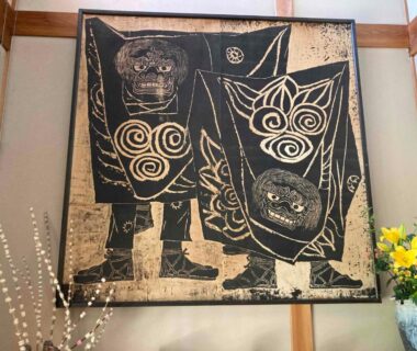 Traditional textile wall hanging with two stylized figures in ryokan interior, wooden beams and floral arrangement visible