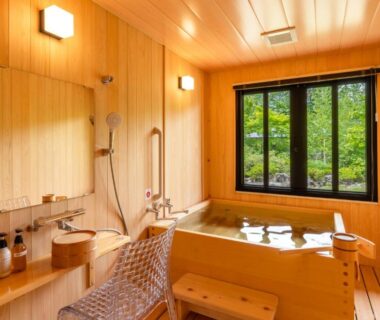 Wooden soaking tub room with window overlooking greenery, shower, wooden stool and mirror at Ryoan Kawaki in Nagano