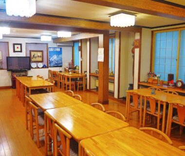 Communal dining room with wooden tables, chairs, shoji screens and overhead lights at 旅館FURUYA in Nagano