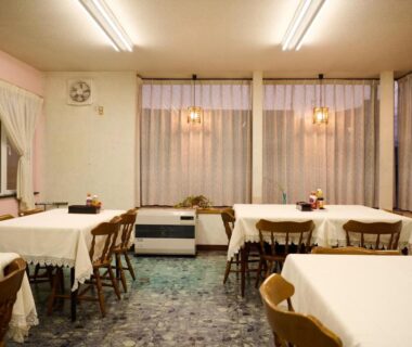 Dining room with white tablecloths, wooden chairs, sheer curtains and soft overhead lighting