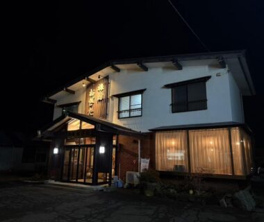 Lit two-story Ryokan Ogita exterior at night with wooden entrance and large curtained windows in Hokkaidō