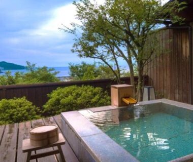 Stone onsen bath on wooden deck with stool, overlooking a wooded cove and bay