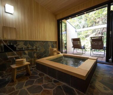Wooden-framed indoor onsen bath with stone floor, stool and open terrace seating overlooking greenery at Yudono Totonoyu Shizuoka