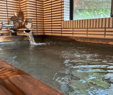 Wooden indoor onsen tub with flowing spout, tiled walls, window and bamboo screen