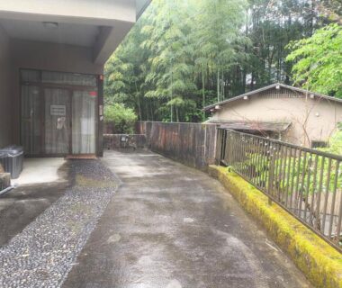 Wet concrete driveway leading to Amakawa Ryokan entrance with bamboo grove and moss-covered low wall