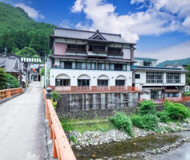 Multi-story ryokan building beside river with bridge and forested mountain backdrop in Dorogawa Onsen, Nara