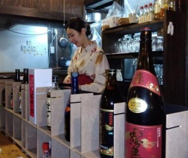 Attendant in kimono behind wooden bar with rows of sake bottles and cups at Onsenya Hirosuke, Fukushima