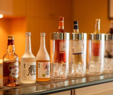 Assorted sake and whisky bottles lined on a wooden bar counter with glassware and warm ambient lighting