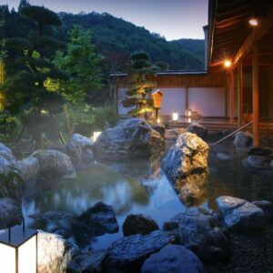 Stone-lined outdoor hot spring with wooden veranda, lanterns, and mountain backdrop at Yūsen Shidate, Iwate, Tōhoku