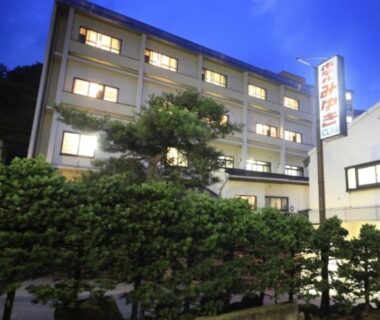 Three-story ryokan building at dusk with illuminated windows, hedge and pine trees in front, signboard visible, Gunma setting