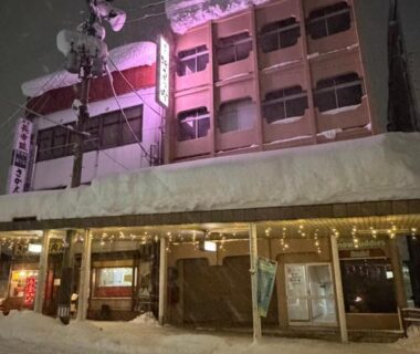 Night exterior of snow-laden ryokan Auberge de poudreuse de Yuzawa with lit windows and covered wooden entrance, Niigata