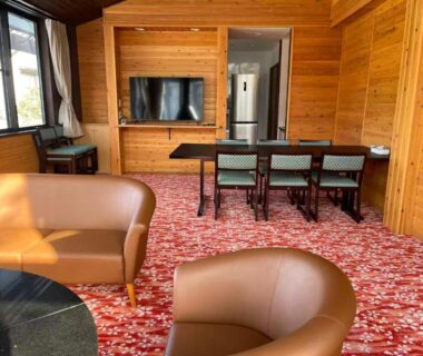 Spacious wood-paneled ryokan lounge with dining table, leather chairs, television and patterned carpet in Gunma