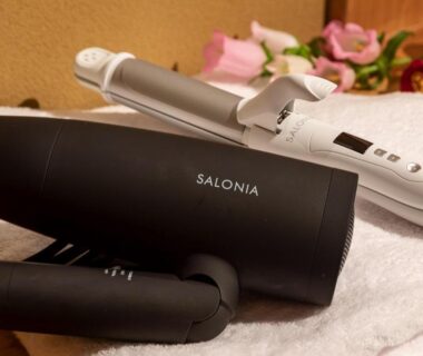 Black hair dryer and white curling iron on folded towel with robe and decorative flowers in guest room