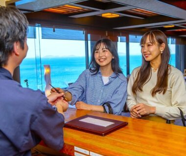 Two women receiving keys at wooden front desk with sea visible through large windows