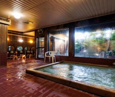 Indoor bathing area with rectangular wooden tub, tiled floor, seating and large window overlooking garden at Ryokufuen Kiyoharu