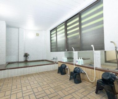 Communal indoor bath with tiled floor, low stools, handheld showers and a soaking tub at Naeba Lodge Oka Niigata