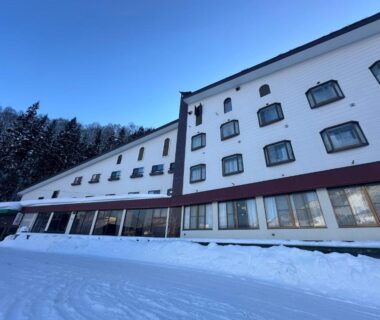 Snow-covered multi-story ryokan exterior with rows of windows and forested slope backdrop in Niigata