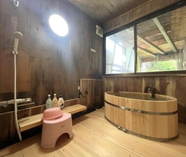 Rustic wooden bathing room with tub, shower area, small stool and garden view at Suzumenoyado Yagura Nagano