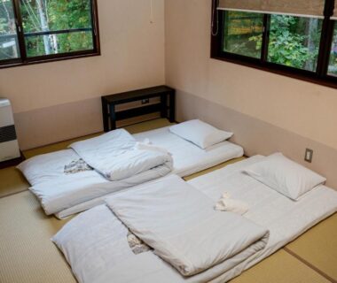 Three futon mattresses on tatami floor in ryokan room with sliding wooden windows overlooking greenery