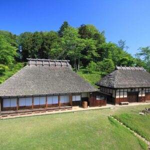 Two thatched roof buildings with wooden facades and grassy courtyard under clear blue sky in Miyagi, Tōhoku