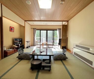Spacious tatami room with low table, floor cushions, sliding balcony doors overlooking wooded area at Kagetsu Highland Hotel, Takayu Onsen