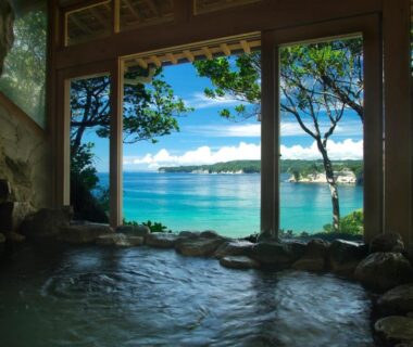 Indoor onsen bath with large window overlooking blue sea and coastal trees at 鵜原館, Chiba