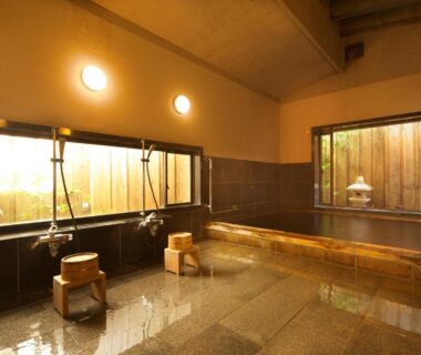 Interior onsen bathing area with wooden soaking tub, tiled floor, stools and small courtyard window